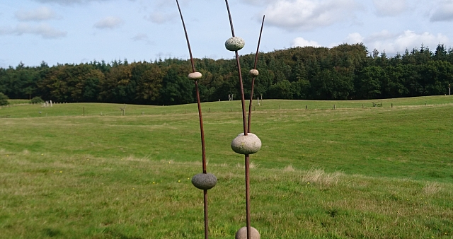 Moderne Skulptur mit Steinen auf Metallstangen in einer Wiesenlandschaft.